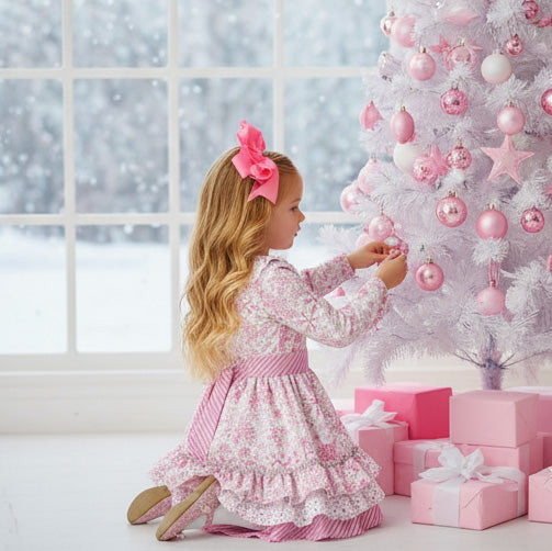 Young girl in a pink snowflake print dress sitting next to a decorated Christmas tree with pink ornaments.