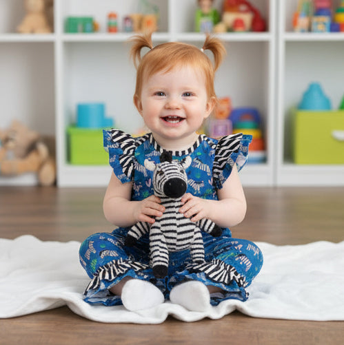 Child wearing a blue romper with zebra patterns holding a stuffed zebra toy in a room with toys in the background.
