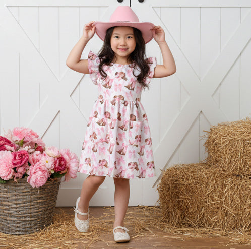 Young girl in a pink cowboy hat print dress and hat standing on straw with flowers and hay bale in the background.
