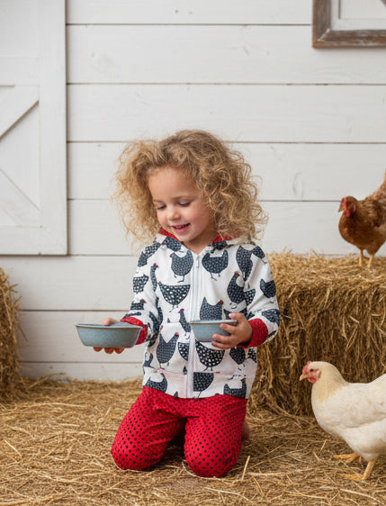 Child in a barn with chickens and hay, wearing a patterned jacket and red pants.