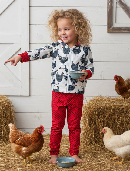 Child in a barn with chickens and hay, wearing a patterned jacket and red pants.