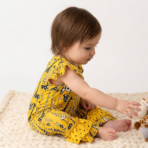 A toddler sitting on a cream knit blanket wearing a yellow sleeveless romper with a black and white animal print, reaching for a plush toy.