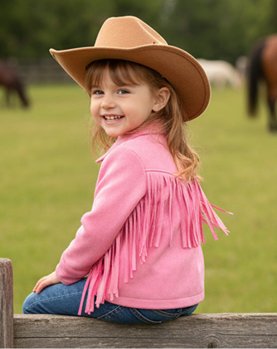 Child wearing a pink fringed jacket and brown cowboy hat sitting on a wooden fence with horses in the background.