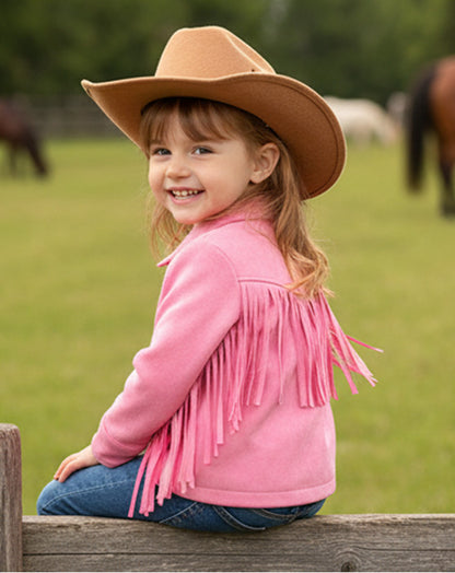 Child wearing a pink fringed jacket and brown cowboy hat sitting on a wooden fence with horses in the background.