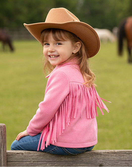 Child wearing a pink fringed jacket and brown cowboy hat sitting on a wooden fence with horses in the background.