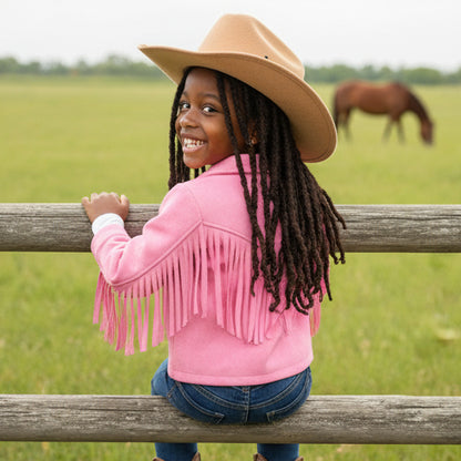 Child wearing a pink fringed jacket and tan cowboy hat standing in a field with a horse in the background.