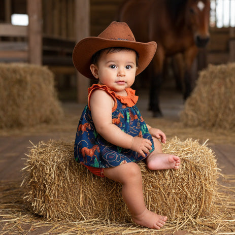 Baby in a cowboy hat sitting on a hay bale with a horse in the background
