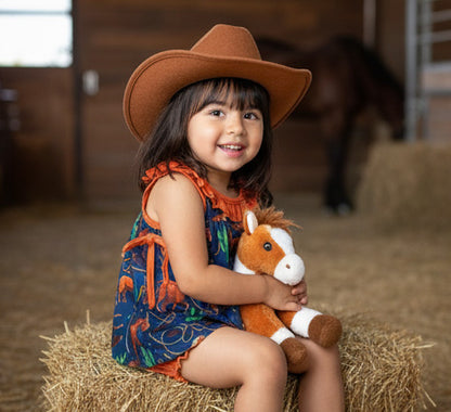 Child wearing a cowboy hat and holding a stuffed horse on a hay bale in a barn setting