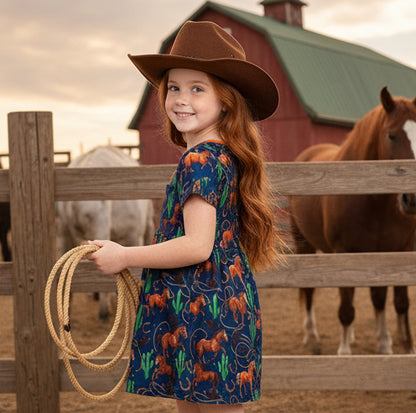 Young girl in a cowboy hat and horse-patterned dress holding a lasso, with a horse and barn in the background.