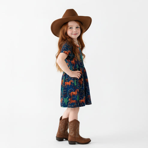 Young girl wearing a horse print patterned dress, cowboy hat, and boots on a white background