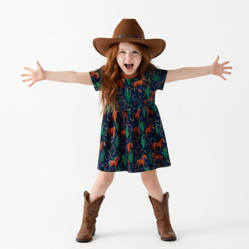 Young girl wearing a horse print patterned dress, cowboy hat, and boots on a white background