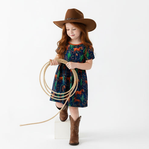 Young girl wearing a horse print patterned dress, cowboy hat, and boots on a white background