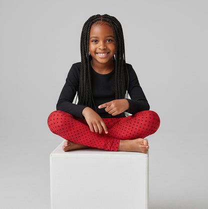 Child wearing black long-sleeve top and red polka dot pants sitting on a white block against a gray background