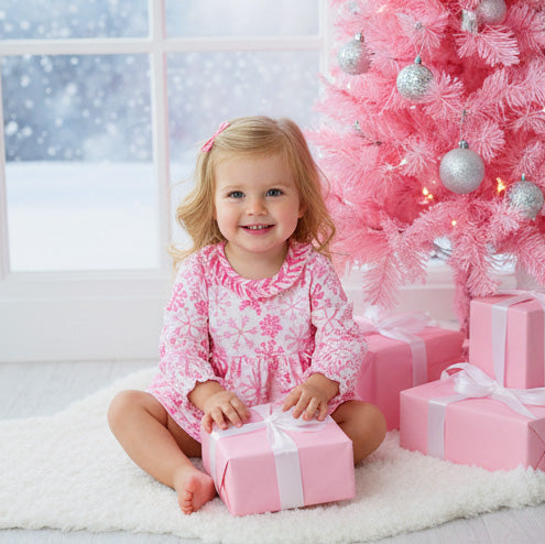 Child in a pink dress sitting next to a Christmas tree with pink decorations and presents.