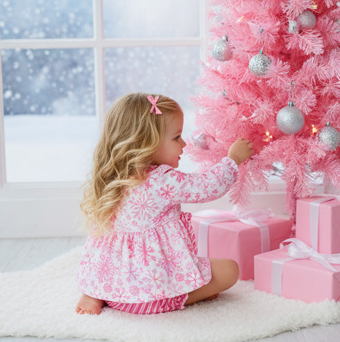 Child in a pink and white dress decorating a pink Christmas tree with silver ornaments.