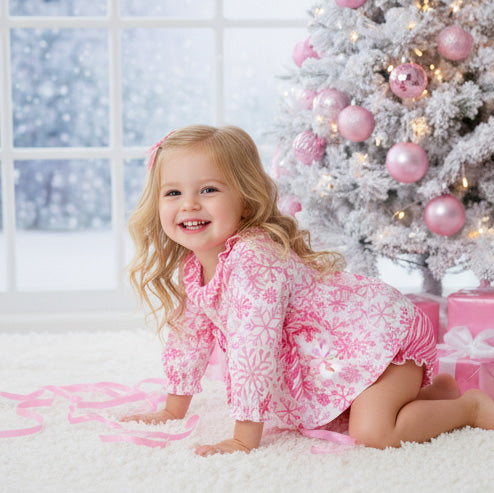 Child in pink snowflake print outfit sitting in front of a decorated Christmas tree with pink ornaments.