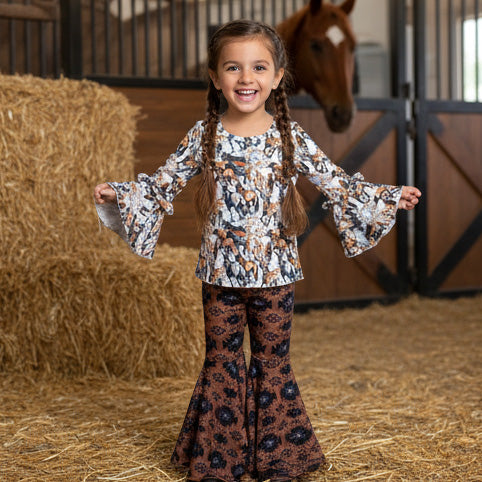 Young girl in a patterned outfit standing in a barn with hay and a horse in the background
