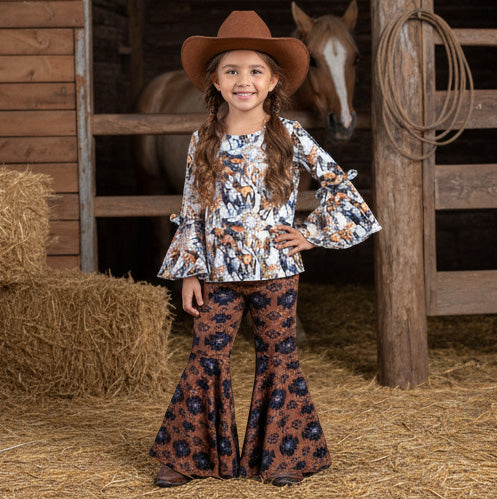 Young girl in a horse print outfit with a cowboy hat standing in a barn with a horse in the background.