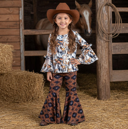 Young girl in a horse print outfit with a cowboy hat standing in a barn with a horse in the background.
