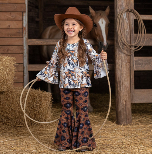 Young girl in a cowboy hat and patterned outfit standing in a barn with a horse and hay bales.