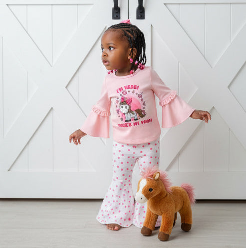 Child wearing a pink outfit with a horse design, standing next to a plush horse toy against a white wooden background.