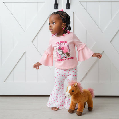 Child wearing a pink outfit with a horse design, standing next to a plush horse toy against a white wooden background.