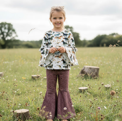 Young girl in a field wearing a patterned top and purple pants.