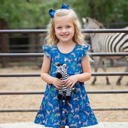 Young girl in a blue dress with zebra pattern holding a stuffed zebra toy, standing in front of a zebra at a zoo.